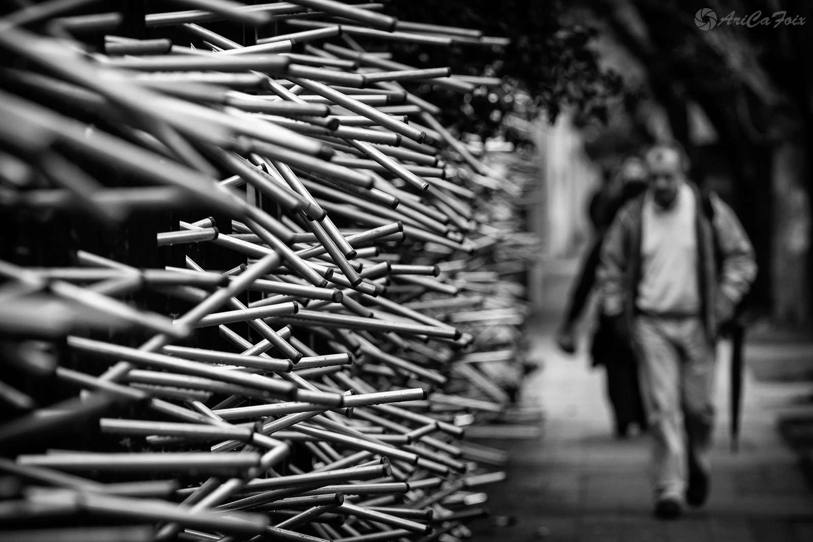 La figura de un hombre camina junto a un muro de sillas cuyas patas apuntan como espinas.